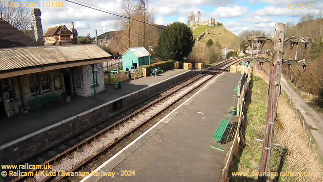 A railway station scene under a partly cloudy sky. On the left, there's a large stone building with a steep roof and wooden details. The platform is lined with green benches and has a sign indicating "WAY OUT." A narrow railway track runs along the front, with another track visible to the right. In the background, a hill rises with a castle on top, surrounded by trees, and additional buildings are visible nearby. A wooden telephone pole stands to the right of the image.