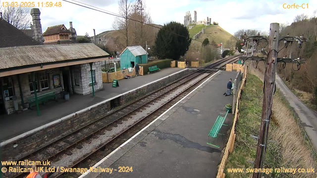 A railway station platform is shown with a wooden shelter on the left, featuring a peaked roof. Green benches are placed along the platform and some in the background. A person is kneeling on the ground near the benches. In the background, there is a green building, and higher up, a hill with remnants of a castle ruins. The sky is partly cloudy, creating a bright but overcast atmosphere. The tracks run alongside the platform, leading to the right side of the image, with a fence and trees in the background.