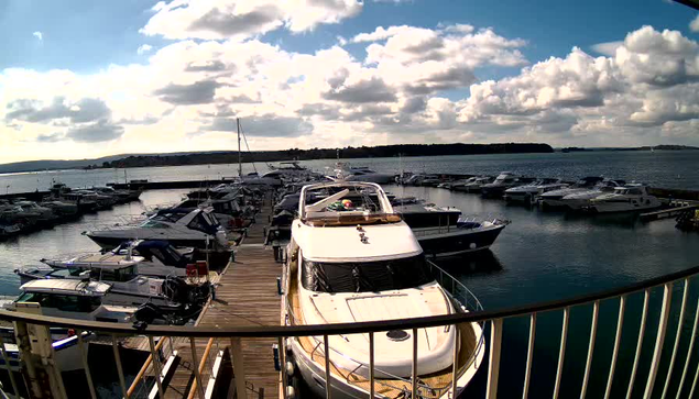 A view from a webcam shows a marina filled with various boats and yachts docked in the water. In the foreground, a larger white yacht is prominent, positioned at the dock. The background features numerous smaller boats, both moored and moving, surrounded by calm water. The sky is partly cloudy with a mix of sunlight and shadows casting over the scene, and a distant landmass is visible on the horizon. The overall atmosphere is tranquil and maritime.