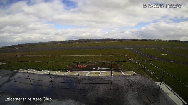 A wide view of an airfield captured from a webcam at Leicestershire Aero Club. The foreground features a rooftop terrace with a railing, and some wet surfaces indicating recent rain. Below, a grassy area leads to a runway, which is mostly empty except for a few marked sections. The sky is partly cloudy with patches of blue peeking through the clouds.