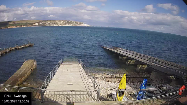 A view of a seaside area featuring calm blue water under a partly cloudy sky. There are two wooden piers extending into the water, one slightly narrower and covered in small railings. On the shore, a concrete ramp leads down to the water's edge, bordered by a rocky beach. A few colorful kayaks, in yellow and blue, are stored near the water on a concrete platform. The landscape is hilly in the background, with white chalk cliffs visible along the coastline. The scene is serene, with a few clouds scattered in the sky.