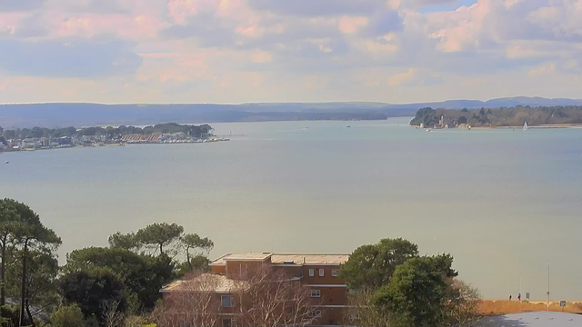 A scenic view of a calm waterway stretches across the image, with gentle waves reflecting the soft colors of the sky. In the distance, a shoreline with various buildings, including a marina, can be seen. Several boats are visible on the water, and lush green trees frame the foreground, adding to the natural beauty. The sky is partly cloudy, suggesting a peaceful daytime atmosphere.