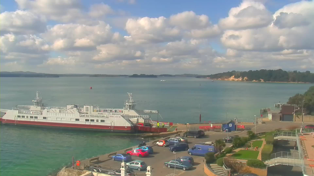 A ferry is docked at a harbor with a calm blue sea in the background. The sky is partly cloudy with large, fluffy clouds. Various cars are parked along the waterfront, including blue and red vehicles. In the foreground, there are flower beds and greenery along a pathway leading to the water. The shoreline features trees and cliffs in the distance.