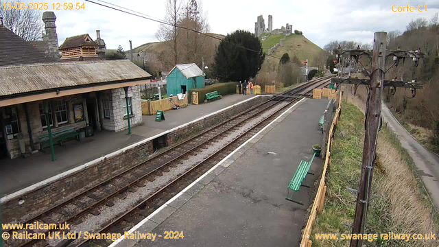 A railway station platform is depicted, featuring green benches along the edge. To the left, there is a stone building with a sloped roof and tall chimney. In the background, a hill rises with the ruins of a castle. A group of four people stands near a fenced area to the left, while a sign indicating "WAY OUT" is positioned on the platform. The tracks extend towards the right side of the image. The overall scene is set on a cloudy day.