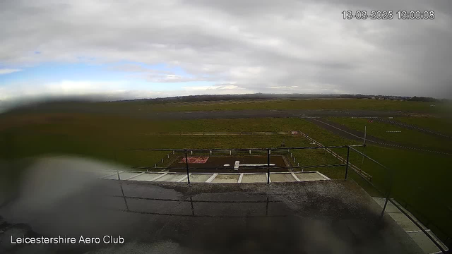 A view from a webcam showing an open grassy field and runway at Leicestershire Aero Club. The sky is mostly cloudy with some patches of blue visible. In the foreground, there is a railing and a wet surface, suggesting recent rain. The field extends into the distance, with a faint outline of hills on the horizon.