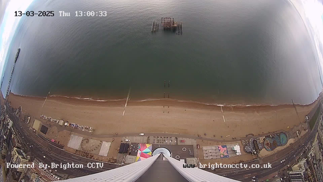 A view from a high vantage point overlooking a sandy beach and the sea. The beach is lined with a pier extending into the water, and several structures and people can be seen along the shore. The area appears to be a busy seaside location, with colorful attractions visible. The ocean is calm, with small waves gently lapping at the shore. In the foreground, there are various buildings, including entertainment venues, and a parking area. The scene captures a cloudy sky.