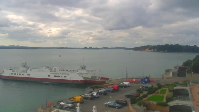 A cloudy sky hovers over a harbor scene. A large white and red ferry is docked at the pier, with a sign indicating "GIVE WAY TO CHAIN FERRY." In the foreground, several parked vehicles of various colors are lined up in a parking lot. To the right, there are green landscaped areas, and the water reflects the overcast sky. In the background, a forested island and gentle waves of the water extend toward the horizon.