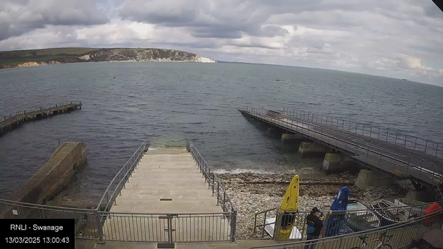 A view of a coastal area featuring a rocky shoreline and calm water. In the foreground, there are two kayak-shaped boats, one yellow and one blue, positioned on a concrete dock. A set of stairs leads down to the water. To the right, a larger pier extends out into the water, with several people visible on it. In the background, a line of cliffs can be seen under a cloudy sky. The scene conveys a tranquil seaside atmosphere.