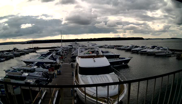 A marina scene featuring numerous boats moored at a dock. In the foreground, a large white yacht is prominent, with a deck and cabin visible. The water reflects a cloudy sky, which is filled with gray clouds and a hint of sunlight peeking through. Several other boats of various sizes are arranged along the dock and on the water, with a distant shoreline visible in the background.