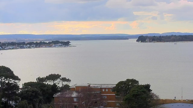 A calm body of water stretches into the distance, reflecting soft light from a cloudy sky. In the foreground, there is a building with multiple windows surrounded by trees. To the left, a coastal area features docks and several boats. The horizon shows a mix of trees and land contours, creating an impression of depth. The overall atmosphere is serene and quiet.