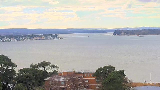 A view of a calm body of water with a range of hills in the background. The sky is partly cloudy with soft colors. In the foreground, there is a cluster of brown buildings surrounded by lush green trees. The shoreline is visible with some boats and buildings along the water's edge. The scene is tranquil and reflects a peaceful atmosphere.