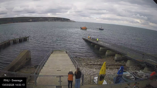 A view of a waterfront scene featuring the sea under a cloudy sky. Two boats are visible on the water, one larger, orange in color, and the other smaller and possibly blue. In the foreground, there is a stone pathway leading down to the water. Two people stand near the edge of this pathway, looking out at the water. On the right, there is a pier with individuals present, and colorful kayaks are seen near the water's edge. The atmosphere appears calm and peaceful.