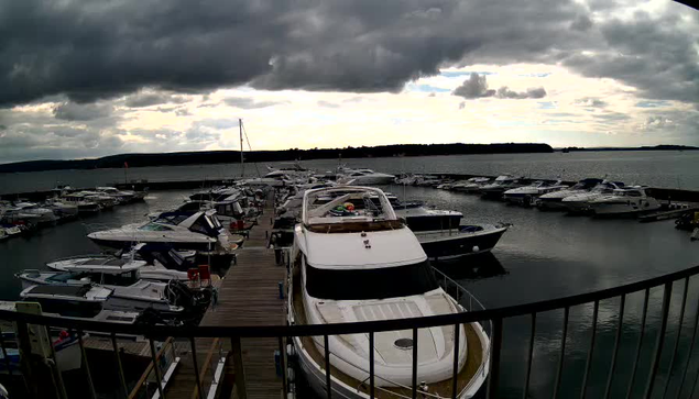 A marina scene with multiple boats docked in calm water. The foreground features various types of boats, including a large white yacht. The background shows a cloudy sky with patches of light peeking through, and a distant shoreline with trees. The image captures a tranquil atmosphere at the marina, with some boats partially obscured by others.