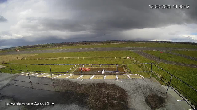 A view from a webcam at Leicestershire Aero Club shows a grassy airfield under a cloudy sky. In the foreground, there is a fenced area with a light-colored surface. The airfield extends into the distance, featuring a runway with markings. The sky is mostly overcast with dark clouds, indicating potential rain. A windsock is visible on the left, showing wind direction.