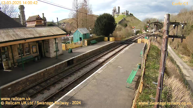 A view of a railway station platform with a stone building on the left featuring a sloped roof and numerous windows. There is a covered bench running along the platform. In the background, there is a green shed and several green benches. To the right, a wooden power pole with wires stands next to the railway tracks. The station is set against a backdrop of a lush, hilly landscape with a castle ruin visible on a hilltop. The sky is partly cloudy.