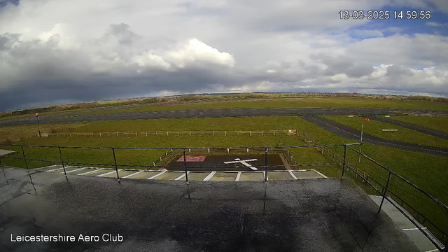 A view from a webcam showing the Leicestershire Aero Club's runway. The foreground includes a marked helipad with a white cross on dark ground, surrounded by grass. In the background, there are sections of runway and taxiway, with a few light clouds in the sky and patches of blue peeking through. The image is taken on a cloudy day with an overall bright yet overcast atmosphere.