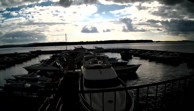 A marina scene showing several boats parked at docks on a calm water surface. The sky is partly cloudy with patches of blue peeking through. The horizon features silhouette-like trees. The overall lighting suggests it is either early morning or late afternoon.