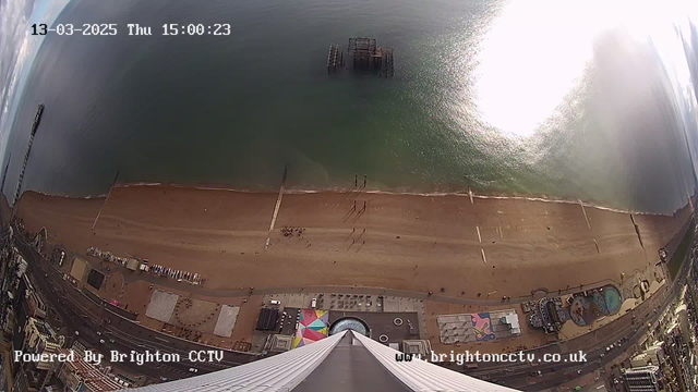 Aerial view of a beach and coastline during daylight. The sandy beach stretches across the lower part of the image, with people walking along it. There is a pier extending into the water, with remnants of an old pier visible halfway submerged. The water is calm and reflects sunlight, creating a shimmer. To the right, a colorful area includes patterns on the ground with a small pool. Buildings and structures line the edge of the beach, with a road running parallel to it. The image is timestamped, indicating the date and time.