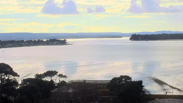 A scenic view of a calm water body under a cloudy sky. The horizon features distant hills and trees along the shoreline, while boats can be seen drifting on the water. In the foreground, the landscape includes a low building surrounded by trees. The colors are soft with a blend of blues, greens, and golden reflections on the water surface, suggesting a tranquil atmosphere.