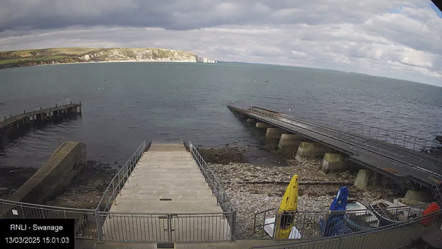 A view of a rocky shoreline and turquoise sea under a partly cloudy sky. In the foreground, a concrete ramp leads down to the water, bordered by a metal railing. To the left, a wooden pier extends into the sea, while on the right, another wooden structure leads further out over the water. Colorful kayaks are stored on the ground, mostly in yellow and blue. The surrounding landscape features green hills in the background, contrasting with the gray clouds overhead.