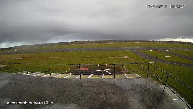 A view from a webcam at Leicestereshire Aero Club shows a grassy airfield under a cloudy sky. In the foreground, there is a tarmac area with a white cross marking a helipad, and a wooden fence surrounds the field. Several strips of asphalt are visible in the distance, with some signs and markers indicating different runway paths. The scene has an overcast sky with varying shades of gray clouds.