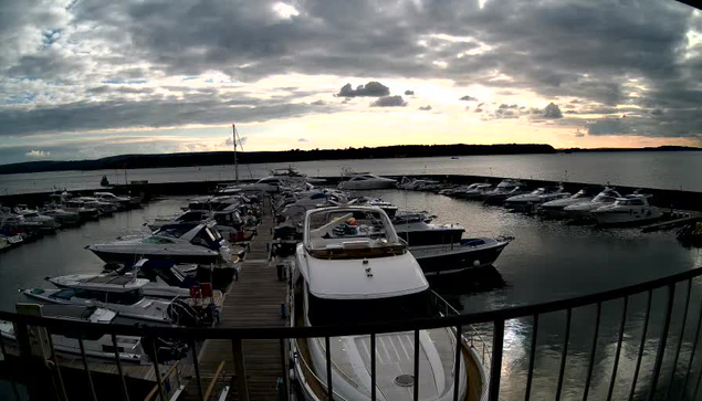 A view of a marina at sunset, featuring numerous boats docked in the water. The foreground shows several boats lined up at the pier, with some larger vessels visible. The sky is partially cloudy with patches of light breaking through, illuminating the calm water. In the distance, hills outline the horizon, creating a serene atmosphere at dusk.