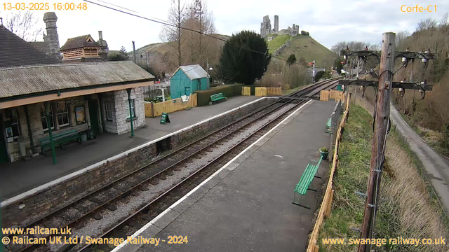 A view of a train station with a stone platform and a thatched roof structure. There are green benches lining the platform, and a small turquoise building can be seen on the left. In the background, a hill rises with the ruins of a castle. The scene is framed by trees and landscaping, with a train track extending towards the horizon. A post with electrical wires and a nearby path are visible to the right. The image appears slightly cloudy, indicating overcast weather.