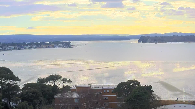 A tranquil view of a calm body of water under a pastel-colored sky, with soft clouds scattered above. In the foreground, there are trees and a building with a flat roof. On the far side of the water, a small town with various structures, possibly boats, can be seen along the shoreline. The distant hills create a gentle backdrop, while the water reflects light, giving it a shimmering effect.