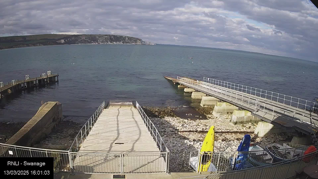 A view of a harbor area with calm blue water and a cloudy sky. On the left, there is a wooden jetty extending into the water. In the foreground, a concrete ramp leads down to a rocky beach. Two colorful kayaks, one yellow and one blue, are displayed on the shore. The background features a rugged coastline with green hills. The image captures a serene seaside atmosphere.