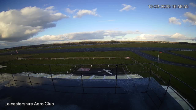 A wide view of an airfield taken from a webcam at Leicestershire Aero Club. The foreground shows a flat rooftop area with a railing, and a painted runway symbol on the ground. In the background, there are grassy fields and a few scattered clouds under a blue sky. The airfield features long stretches of asphalt runway and a fence line separating it from the grass. Overall, the scene depicts an open, aviation-related landscape.