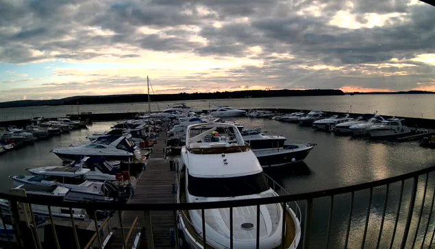 A view of a marina during sunset, with several boats docked in the water. The sky is mostly cloudy, with hints of orange and blue as the sun sets on the horizon. In the foreground, the front of a large white boat is visible, alongside various other boats lined up in the marina. The water is calm, reflecting the colors of the sky. A few distant hills can be seen beyond the water.