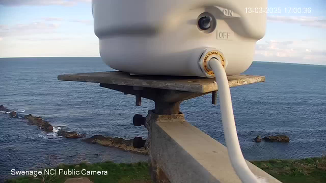 A white device mounted on a metal platform overlooking the ocean. The device has an on/off switch, and a white cable connects it to a surface below. In the background, the calm sea stretches to the horizon under a partly cloudy sky. Rocky formations are visible along the shoreline.