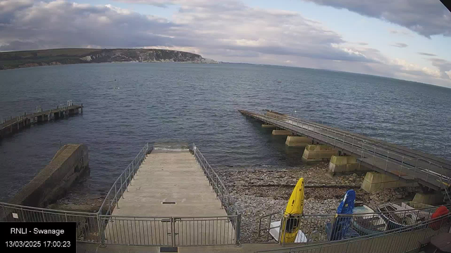 A view of a coastal area featuring the sea, with a rocky shoreline visible. In the foreground, there is a concrete ramp leading down towards the water, with a railing on both sides. To the right, two kayaks are parked on the ground, one yellow and one blue. In the background, a wooden pier extends into the water from the left side. The sky is slightly cloudy, and the coastline with hills stretches in the distance. The time and date are displayed in the bottom left corner.