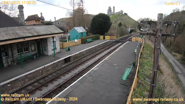 A railway station platform with a stone building featuring a sloped roof and several windows. There are green benches along the platform, and a rustic wooden fence surrounds the area. In the background, a hill is visible with ruins of a castle at the top. The sky is partly cloudy, and railroad tracks run along the platform. A wooden utility pole with wires is seen on the right side of the image. A small turquoise shed is located on the left side of the platform.