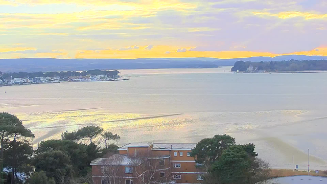 A scenic view of a body of water under a colorful sky at sunset. The foreground features a building with multiple floors and a flat roof, partially obscured by trees. On the opposite shore, there are various structures, possibly boats or piers, silhouetted against the water. The horizon shows distant hills, and the sky is filled with soft clouds illuminated by shades of orange, pink, and yellow. The water's surface reflects the colors of the sky, creating a tranquil, picturesque scene.