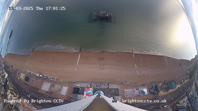 A wide aerial view of a beach with golden sand meeting a calm sea. The shoreline extends across the image, with gentle waves lapping at the edge. In the water, a partially submerged pier structure is visible. The beach is mostly empty, with a few people walking along the edge. Below, there are colorful structures and a circular seating area. The sky above is partly cloudy, and the scene has a serene atmosphere, indicating late afternoon light. The date and time are displayed in the upper left corner.