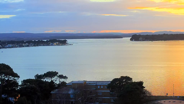 A serene landscape shows a calm body of water reflecting soft shades of blue and orange from the sky. In the distance, a shoreline features a cluster of buildings and boats, while lush green trees line the waterfront. The horizon is marked by gentle hills under a cloudy sky, with the sun setting, casting warm hues across the scene.