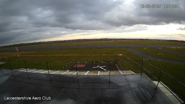 A view from the Leicestershire Aero Club webcam shows an expansive open field with a runway in the distance. The ground is covered with green grass and is mostly clear, with some markings visible. There's a control tower's railing in the foreground, and a faint outline of a cloudy sky can be seen above. The image is timestamped "13-03-2025 18:00:25."