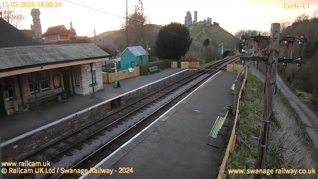 A view of a railway station platform with several benches. In the background, there is a green building and a wooden fence. The tracks run along the bottom of the image, with a grassy hill topped by ruins in the distance. The sky is light as the sun sets, casting a soft glow over the scene. The overall atmosphere is calm and rural.