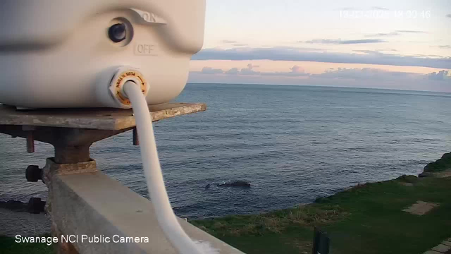 A close-up view of a white device mounted on a metal structure, with a white cable extending from it. In the background, the calm sea is visible under a partly cloudy sky, with soft pastel colors of sunset. The shoreline is grassy with some rocky areas visible in the water. The image includes a timestamp in the upper corner showing the date and time.