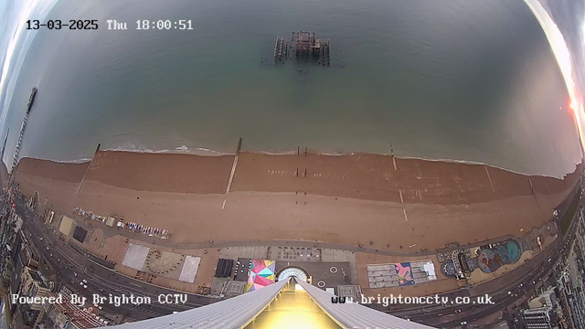 A panoramic view of a beach scene captured by a webcam. The image shows a sandy beach with a few people walking along the shore. There is a pier extending into the sea, and remnants of a dismantled pier in the water. The beach is lined with colorful beach huts and amenities. In the background, the ocean meets the horizon, and the sky features subtle clouds. The timestamp in the top left corner indicates the date and time of the capture.