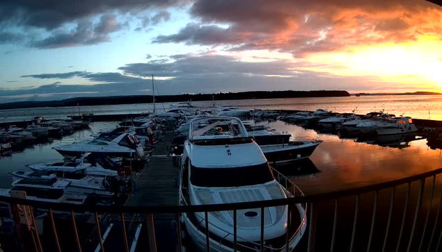 A marina at sunset, featuring several boats docked along a wooden pier. The water reflects the vibrant colors of the sky, with shades of orange, pink, and purple. In the foreground, a larger white boat is visible, with smaller boats around it. The horizon shows distant land and clouds, while the overall atmosphere conveys a peaceful evening scene.