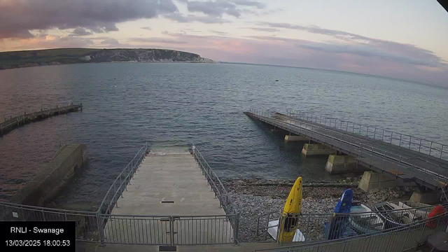 A view of a pier extending into a calm body of water, with a rocky shoreline in the foreground. Two boats, one yellow and one blue, are moored near the edge. There is a concrete ramp leading down to the water, and additional docks visible in the background. The sky is partially cloudy with hints of color, suggesting sunset. In the distance, a hilly coastline is visible. A timestamp and label indicating "RNLI - Swanage" are at the bottom.