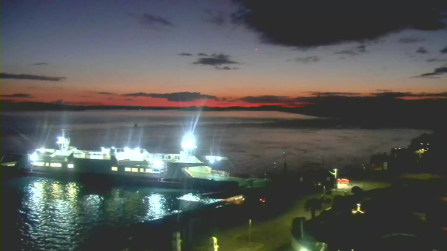 A brightly lit ferry is docked at a harbor during twilight. The ferry's lights reflect on the calm water, with a gradient sky transitioning from orange to dark blue as the sun sets. Silhouetted clouds are scattered across the sky, and faint outlines of land are visible in the distance. The harbor area has some illuminated pathways and structures, adding to the evening ambiance.