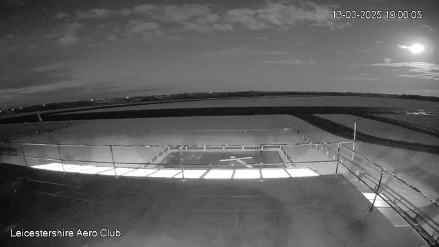 A night view of the Leicestershire Aero Club, with a helipad illuminated below. The scene is predominantly dark, showcasing a clear sky with a few scattered clouds. A bright moon is visible in the upper right corner, casting light over the area. The helipad features a white cross marking. In the background, the horizon is faintly visible against the dark landscape. There are railings along the edge of the structure, and the date and time are displayed in the upper right corner.