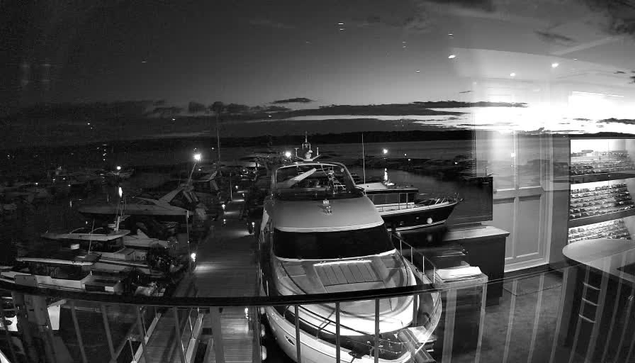 A black and white image showing a marina at dusk. Several boats are lined up in a dock, with the majority being yachts. The water reflects the fading light and the clouds in the sky. There is a railing in the foreground, and the scene gives a sense of calmness and tranquility as night approaches.