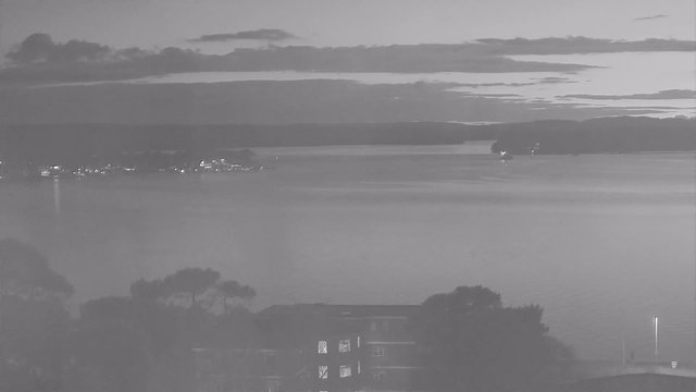 A black and white image shows a calm body of water at dusk. In the foreground, a silhouette of trees can be seen, while a building with illuminated windows is at the bottom center. The middle ground features a quiet waterway reflecting faint light from distant shoreline structures. The horizon is lined with low hills under a cloudy sky, creating a tranquil scene.