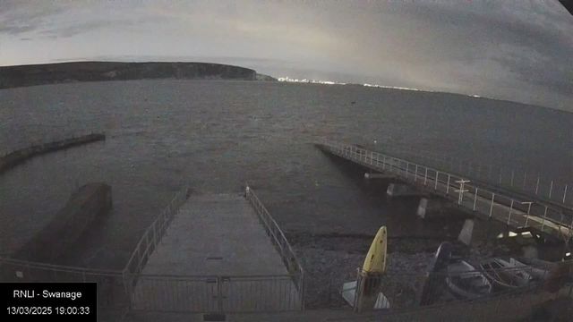 A view of a dark, cloudy evening over a body of water with gentle waves. In the foreground, a concrete pathway leads to a wooden pier extending into the water on the right. To the left, a rocky shoreline is visible, with a yellow kayak and a blue kayak resting on the ground. In the distance, faint city lights are visible along the horizon. The overall atmosphere is calm and serene.