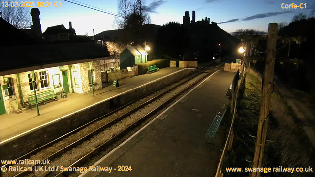 A dimly lit railway platform is visible, featuring benches along the waiting area. There is a green structure with windows, likely a station building, situated to the left. The platform is lined with railroad tracks that stretch into the distance. In the background, there are hills with ruins silhouetted against the evening sky. A single lamp post provides light on the platform, and a sign indicating "WAY OUT" is positioned near the building. The atmosphere suggests early evening, with a hint of twilight in the sky.