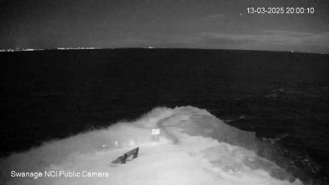 A dark image of the ocean at night, with gentle waves crashing against a rocky shore. A bench is positioned on the edge of the land, and a sign is visible nearby. The horizon features faint lights, possibly from distant buildings or ships. The overall scene conveys a calm and serene atmosphere, with a cloudy sky above.